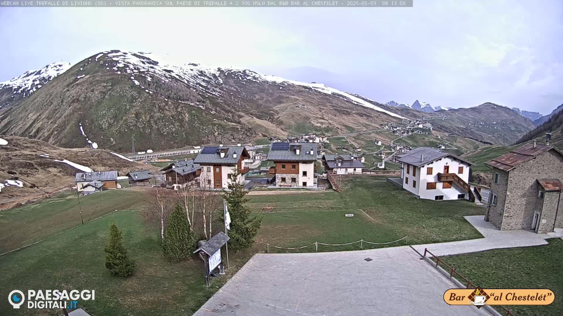 A mountain village nestled in a valley, with snow-capped mountains in the background under a partly cloudy sky.