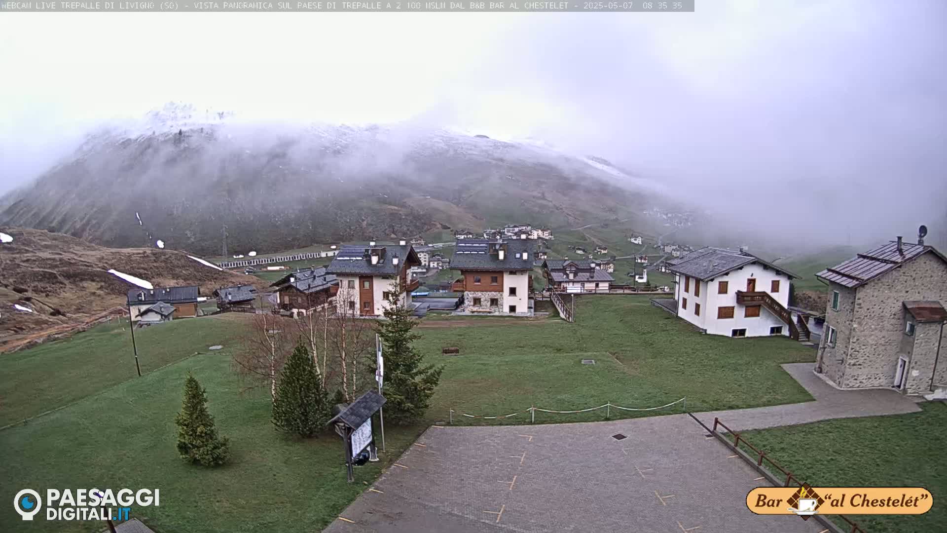 A fog-shrouded mountain overlooks a small village of low buildings and grassy areas.