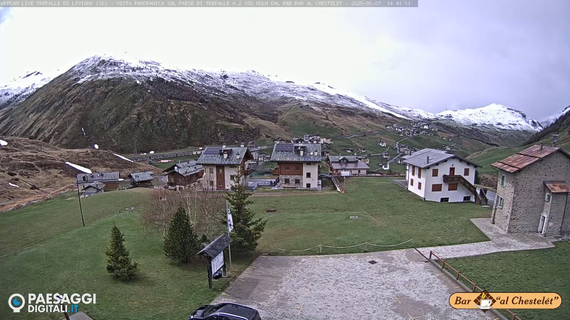 A mountain village with several buildings sits on a grassy hillside under an overcast sky, with snow-capped mountains in the background.