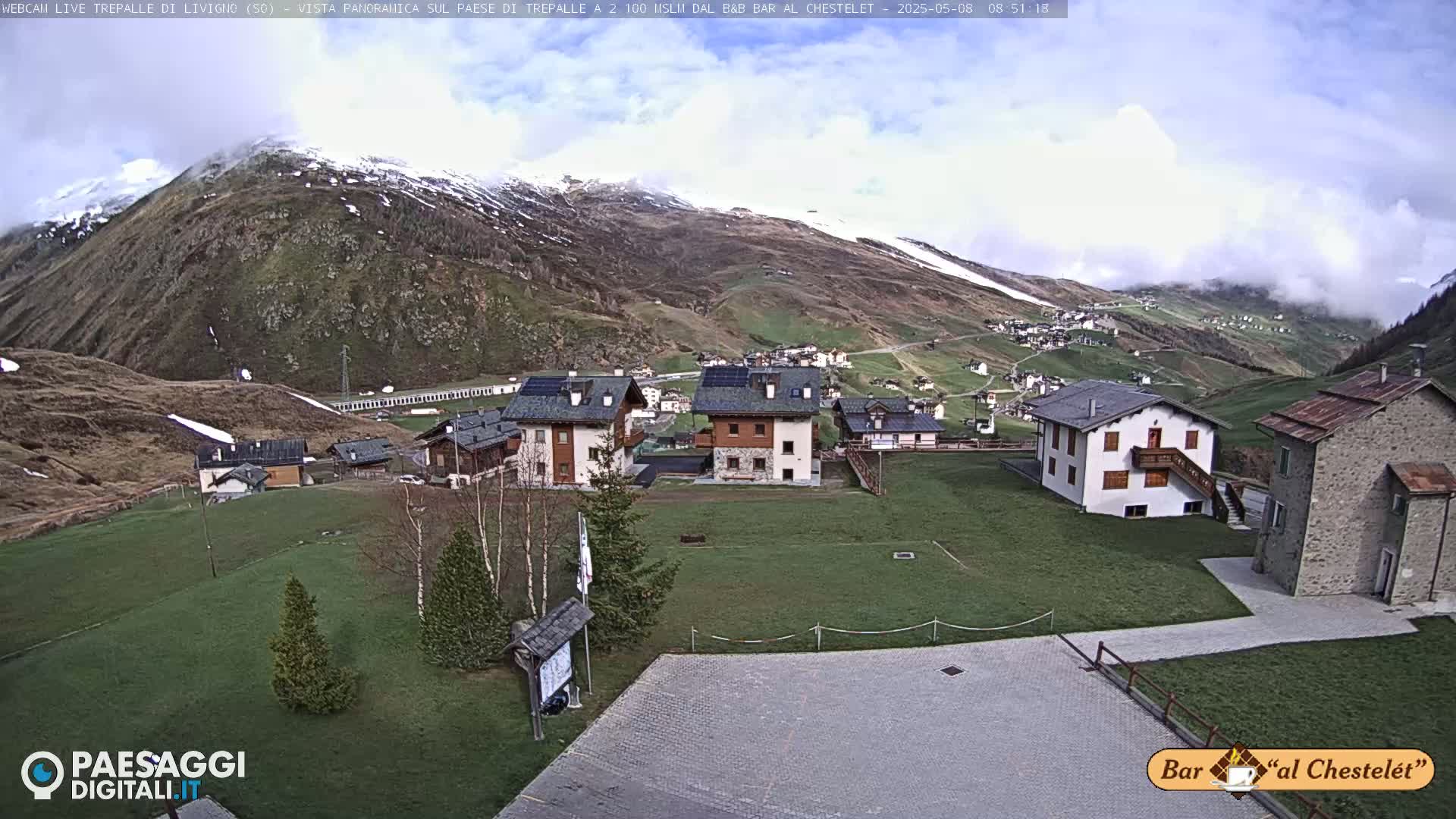 A partly cloudy day shows a small village nestled in a valley, surrounded by green hills and snow-capped mountains.