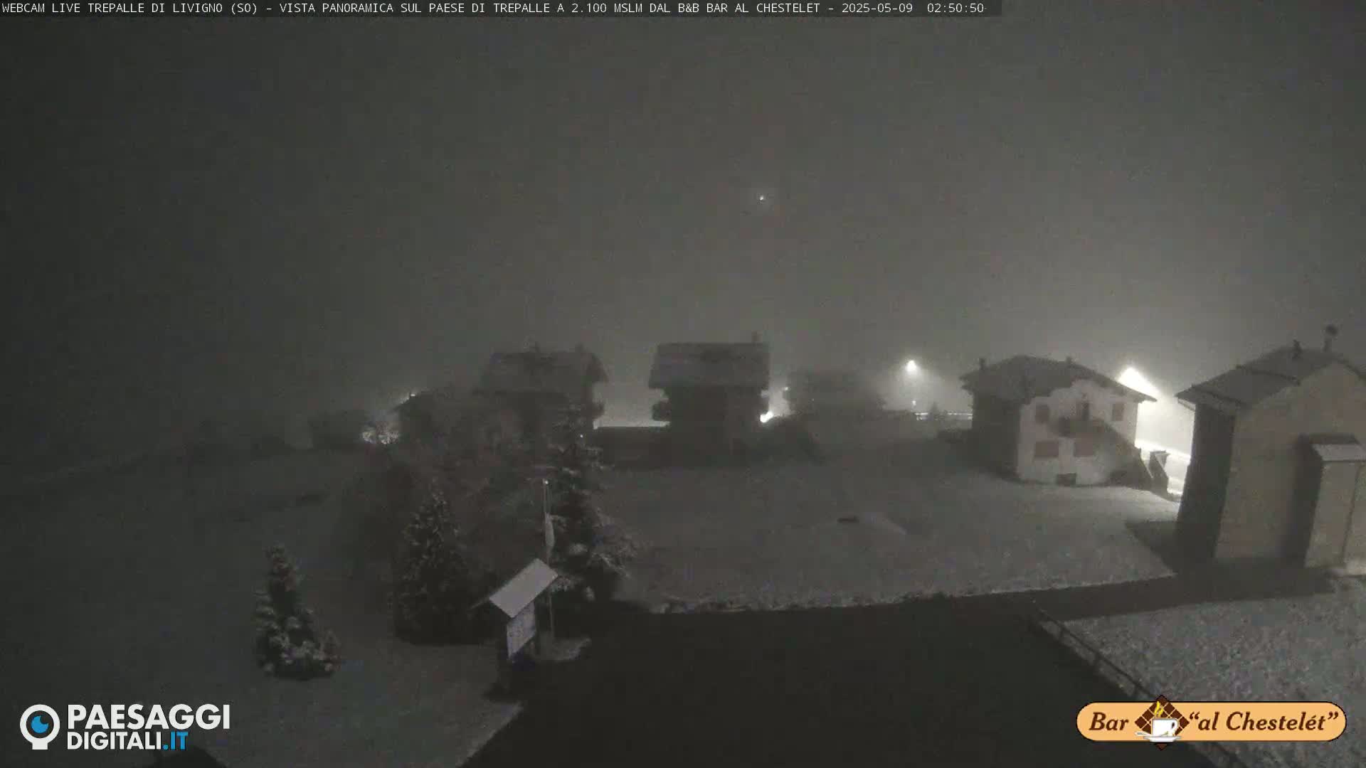 A snow-covered village scene is visible under a dark, foggy sky at night, with several buildings illuminated by streetlights.