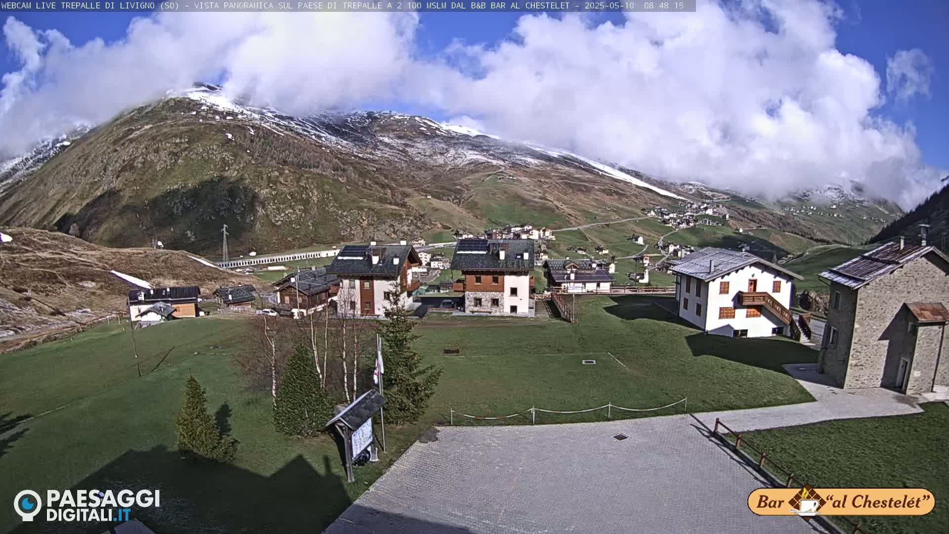 A mountain village nestled in a valley under a partly cloudy sky, with snow visible on the surrounding peaks and green grass in the foreground.