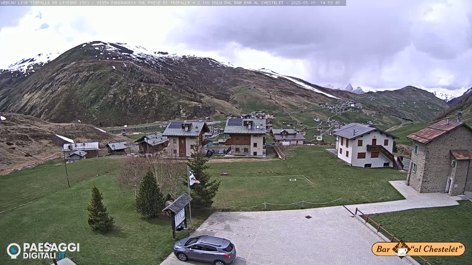 A mountain village with overcast skies and patchy snow on the surrounding hills is viewed from a slightly elevated position, showing a few cars parked in a small lot in the foreground.