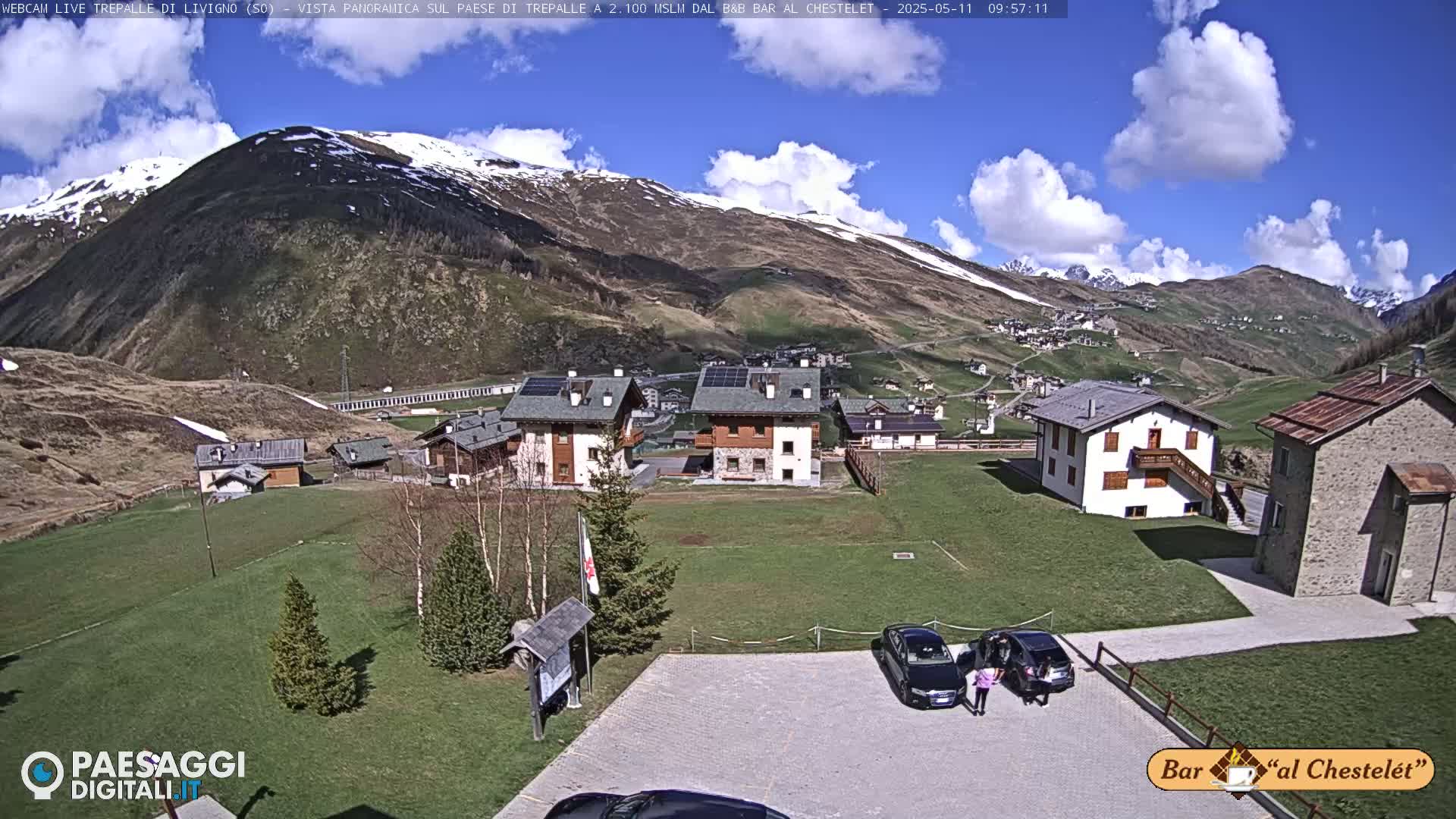 A sunny mountain village with a few cars in a parking lot is nestled below a snow-capped mountain under a partly cloudy sky.