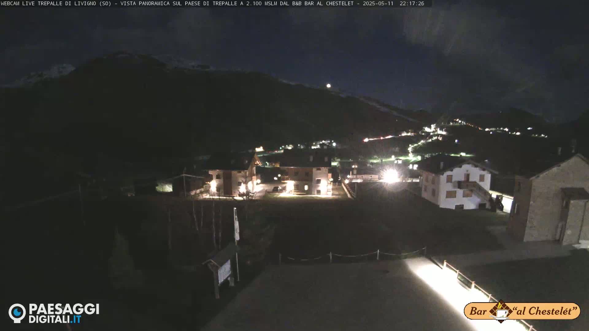 A nighttime, panoramic view of a small village nestled in a mountain valley under a dark sky with a few lights illuminating buildings.