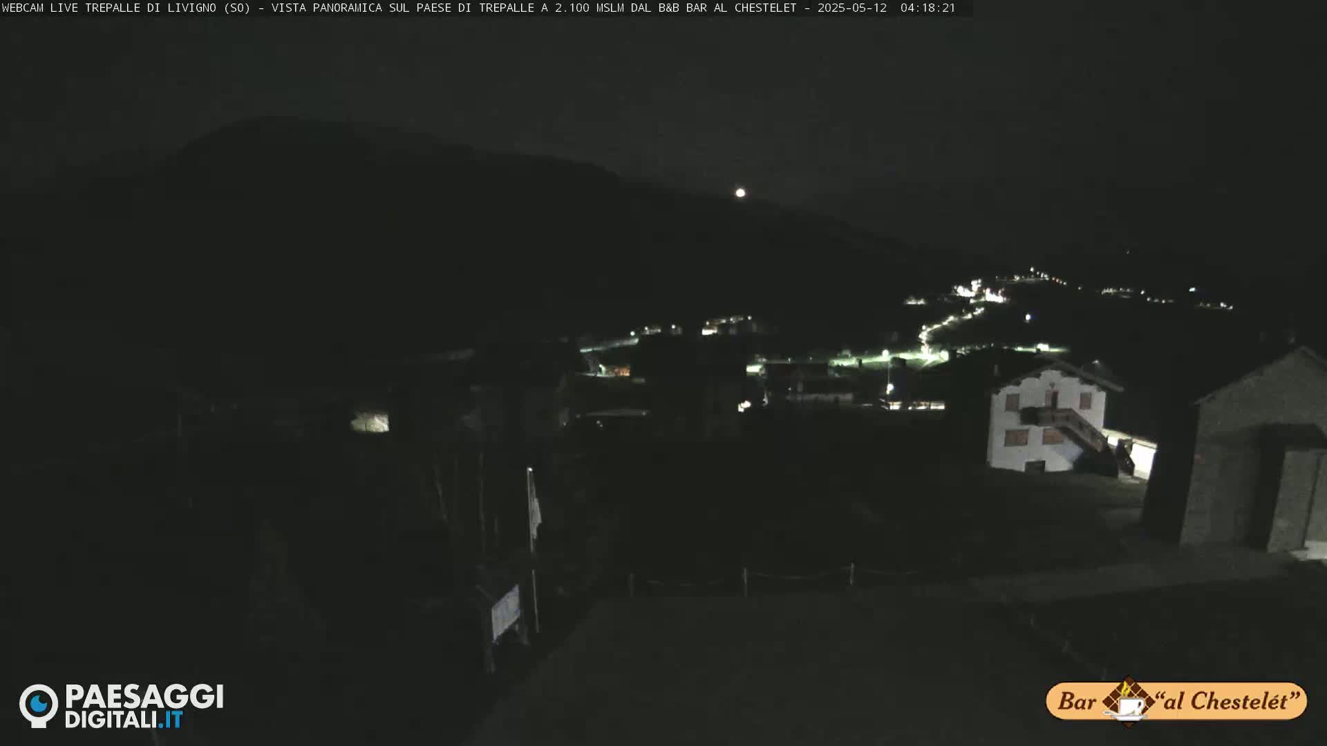 A nighttime, long shot of a small village nestled in a dark mountain valley under a clear sky with a bright moon.
