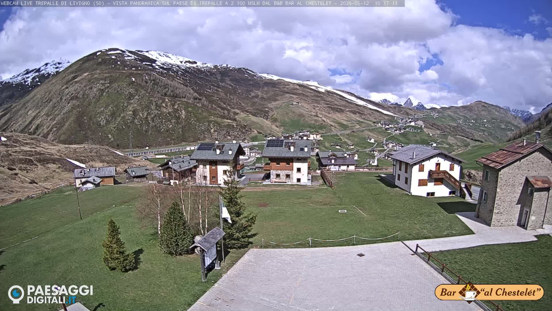 A partly sunny day reveals a village nestled in a valley surrounded by snow-capped mountains.