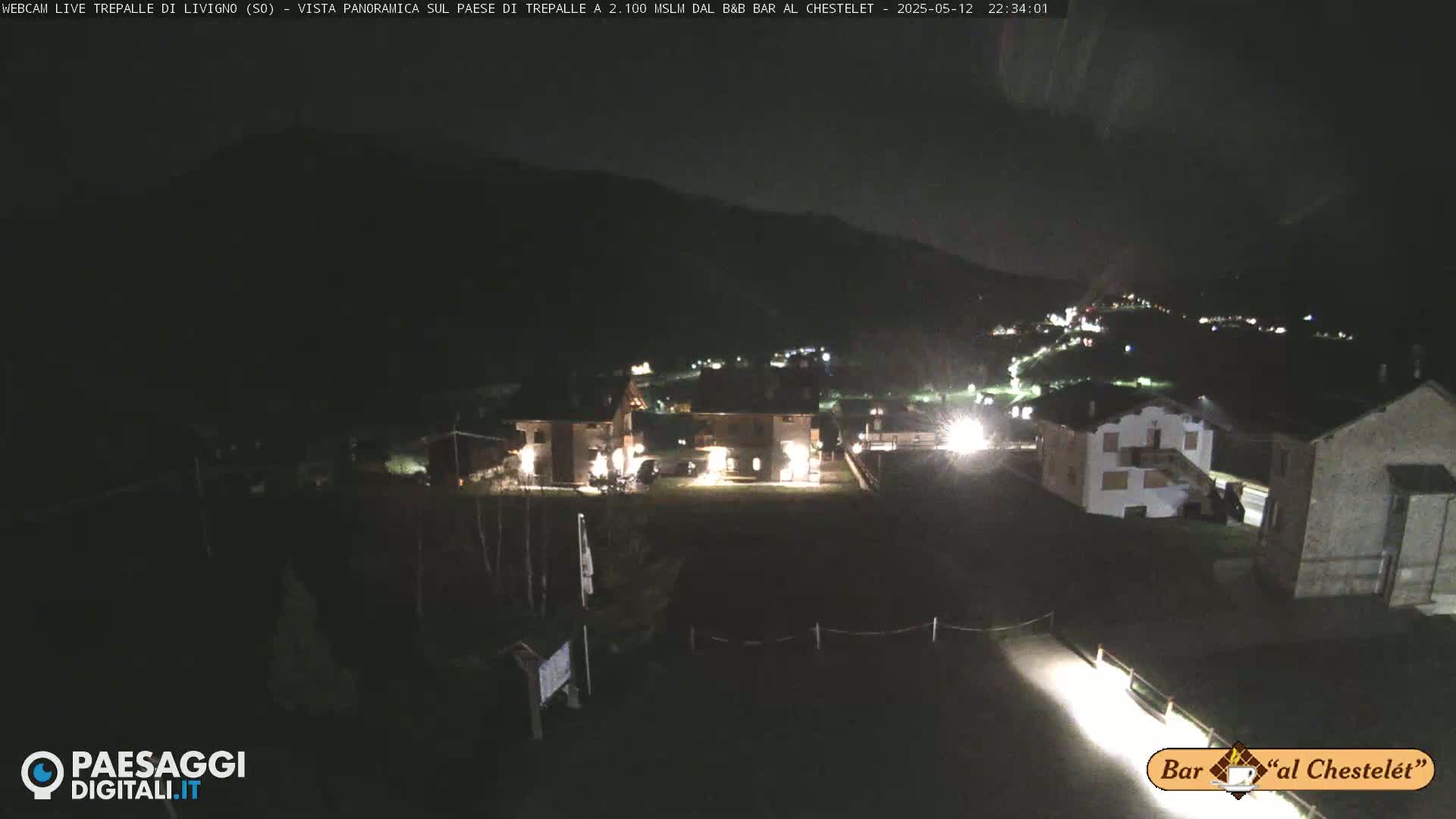 A nighttime view of a small village nestled in a valley, illuminated by scattered lights against a dark mountain backdrop.