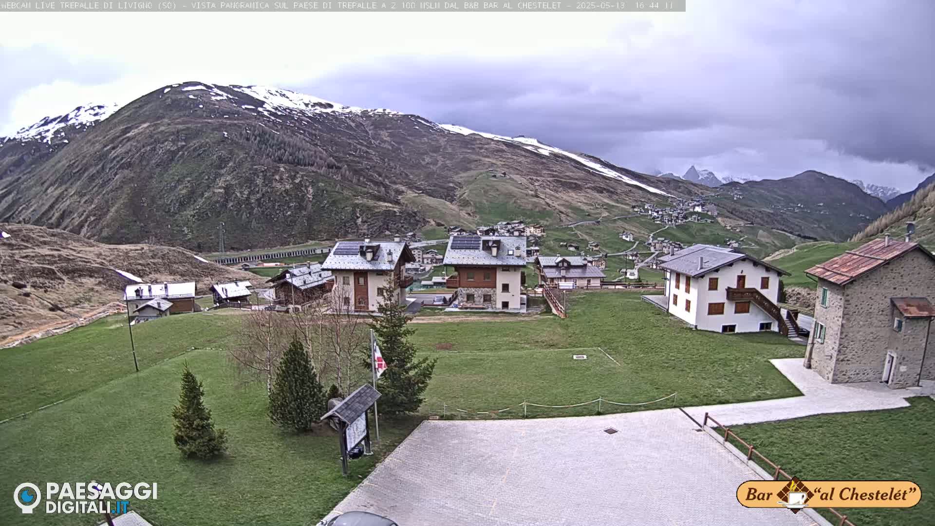 A mountain village with several buildings is situated on a grassy hillside under an overcast sky, with snow-capped mountains visible in the background.