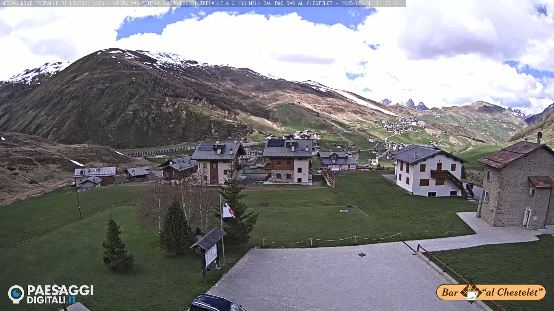A partly cloudy day reveals a small village nestled in a valley surrounded by snow-capped mountains.