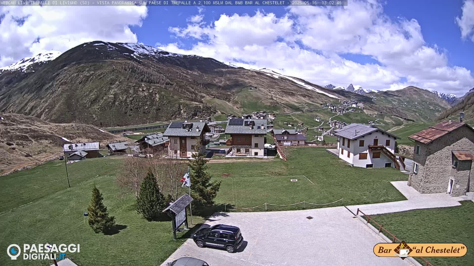 A mountain village with several buildings and a grassy area is seen on a partly cloudy day, with mountains in the background showing some snow.