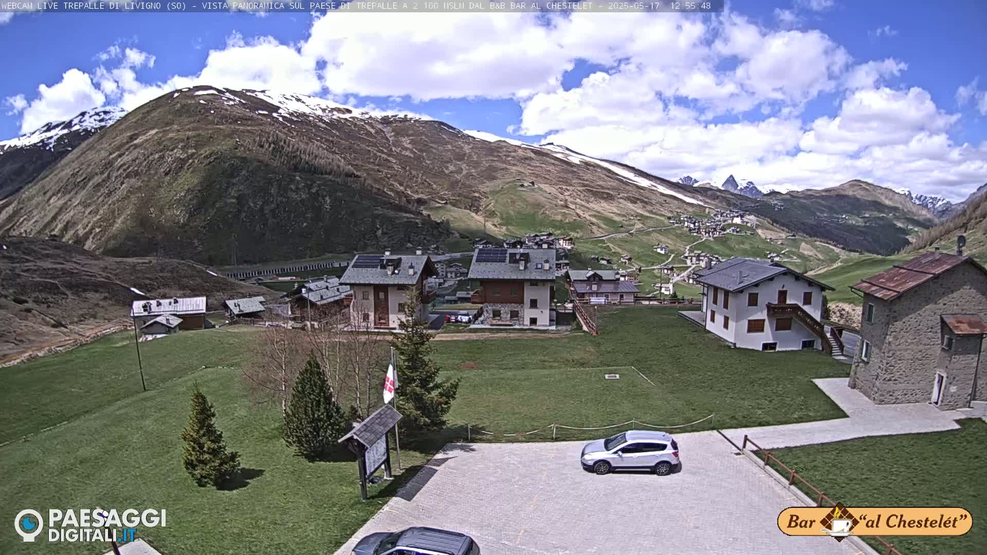 A partly cloudy day reveals a village nestled in a valley surrounded by snow-capped mountains.