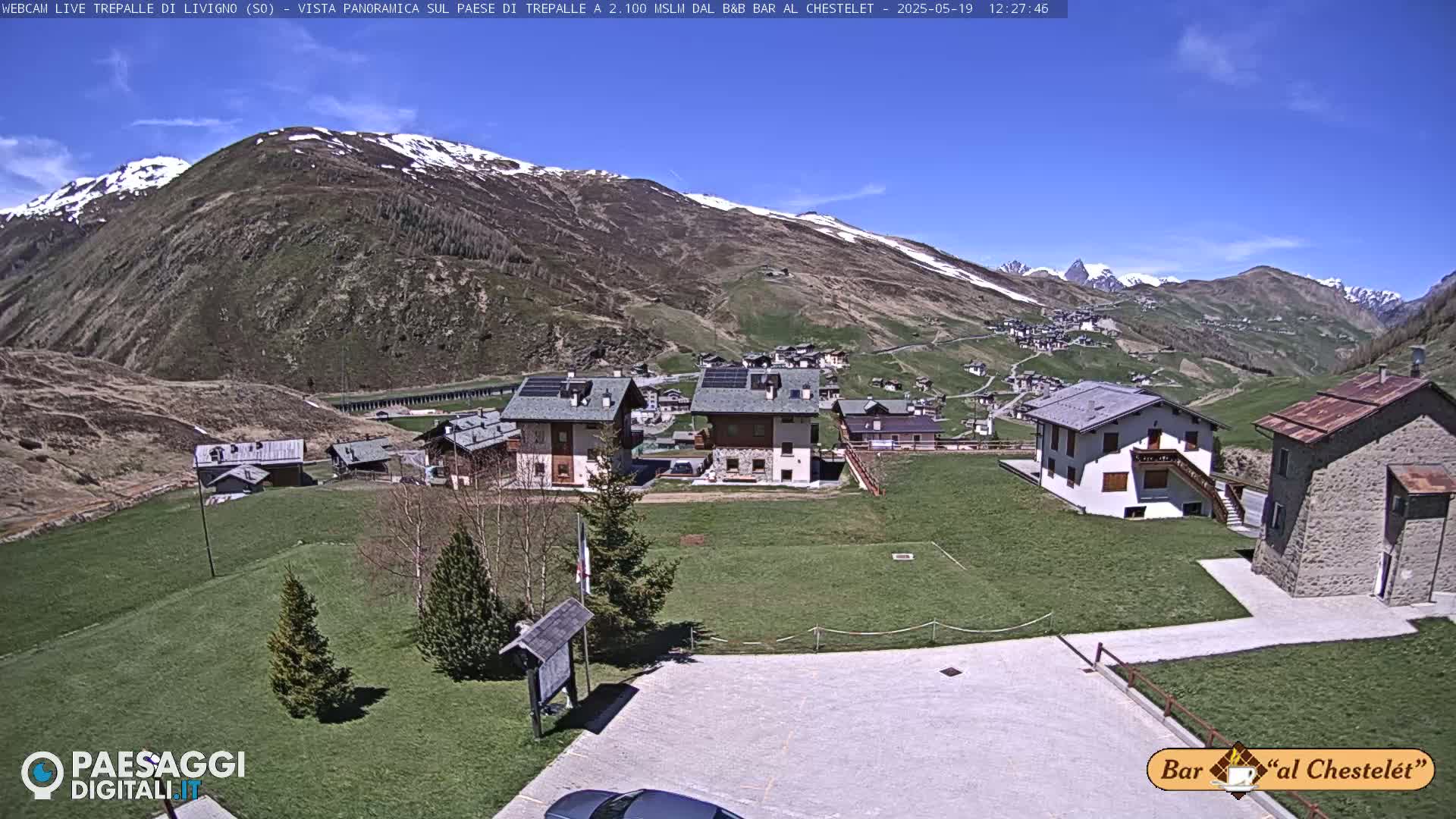 A sunny day reveals a small village nestled in a valley, surrounded by snow-capped mountains and green fields.