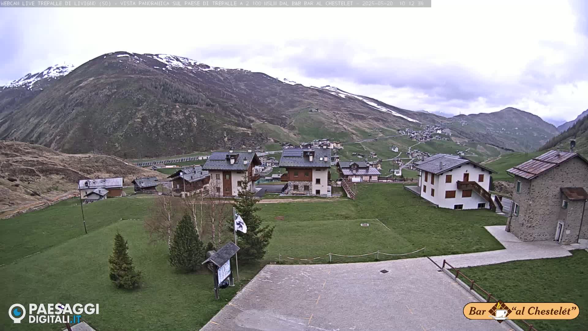 A village nestled in a valley surrounded by mountains with some snow on the higher peaks under a cloudy sky.