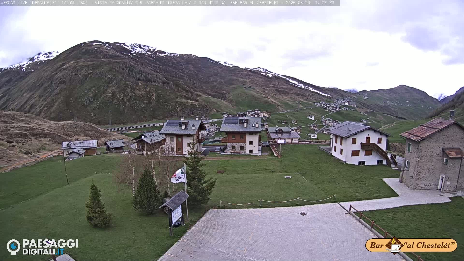 A small village nestled in a valley surrounded by mountains with patches of snow, under a cloudy sky.