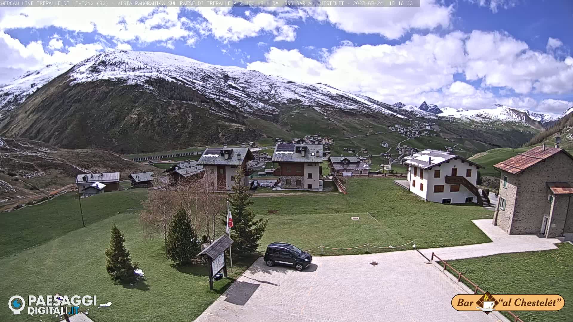 A partly cloudy day reveals a village nestled in a valley, surrounded by snow-capped mountains and green pastures.