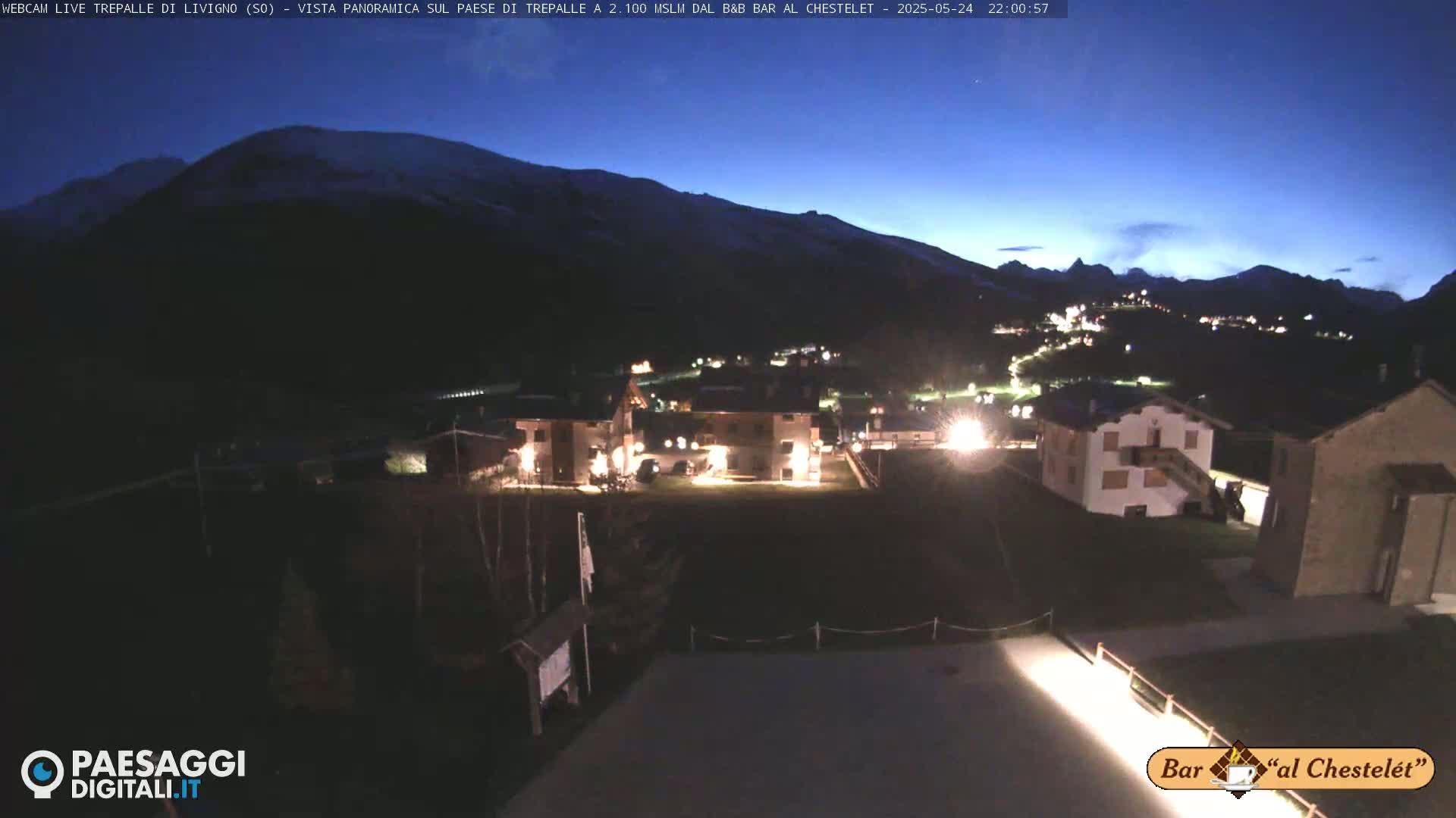 A nighttime view of a snow-capped mountain and a small village illuminated by lights under a dark blue twilight sky.