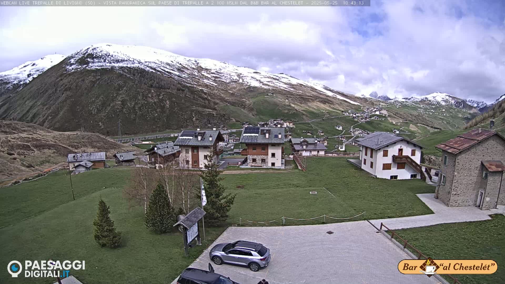A partly cloudy day reveals a snow-dusted mountain overlooking a small village of houses and a grassy area with a few cars parked.