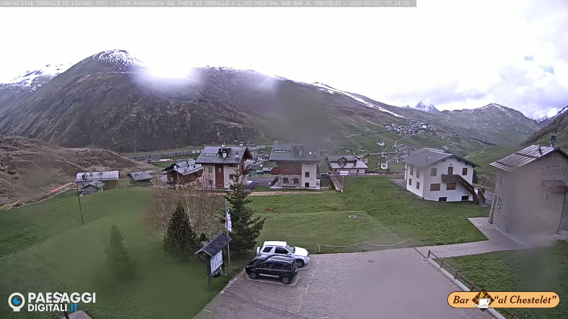 A mountain village with several buildings and two cars parked in a lot, under an overcast sky with some snow visible on the mountain in the background.