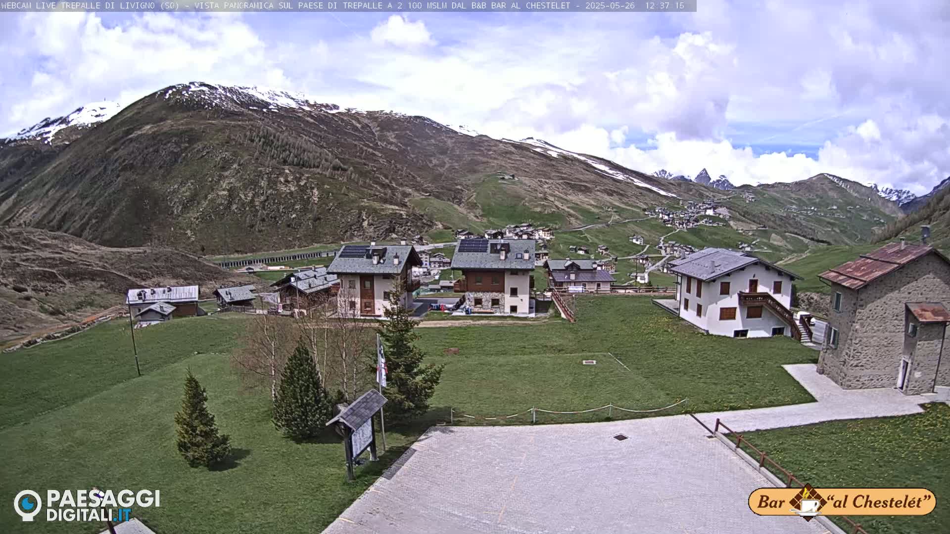 A village nestled in a mountain valley under a partly cloudy sky, with snow still visible on the higher peaks.