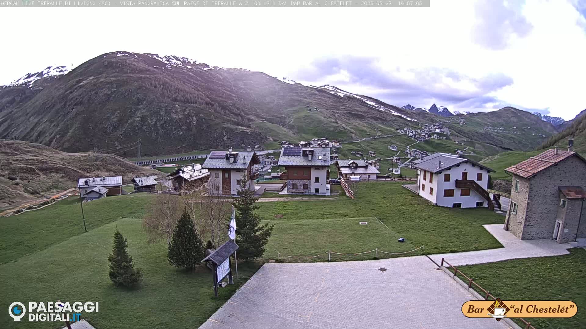 A mountain village with several buildings sits on a grassy hillside beneath a partly cloudy sky, with snow visible on distant mountain peaks.