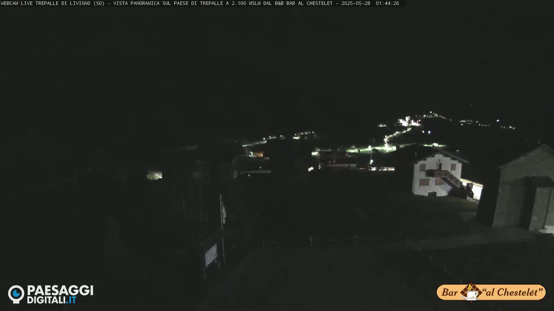 A nighttime, long shot of a village with dimly lit buildings and winding roads under a dark sky.