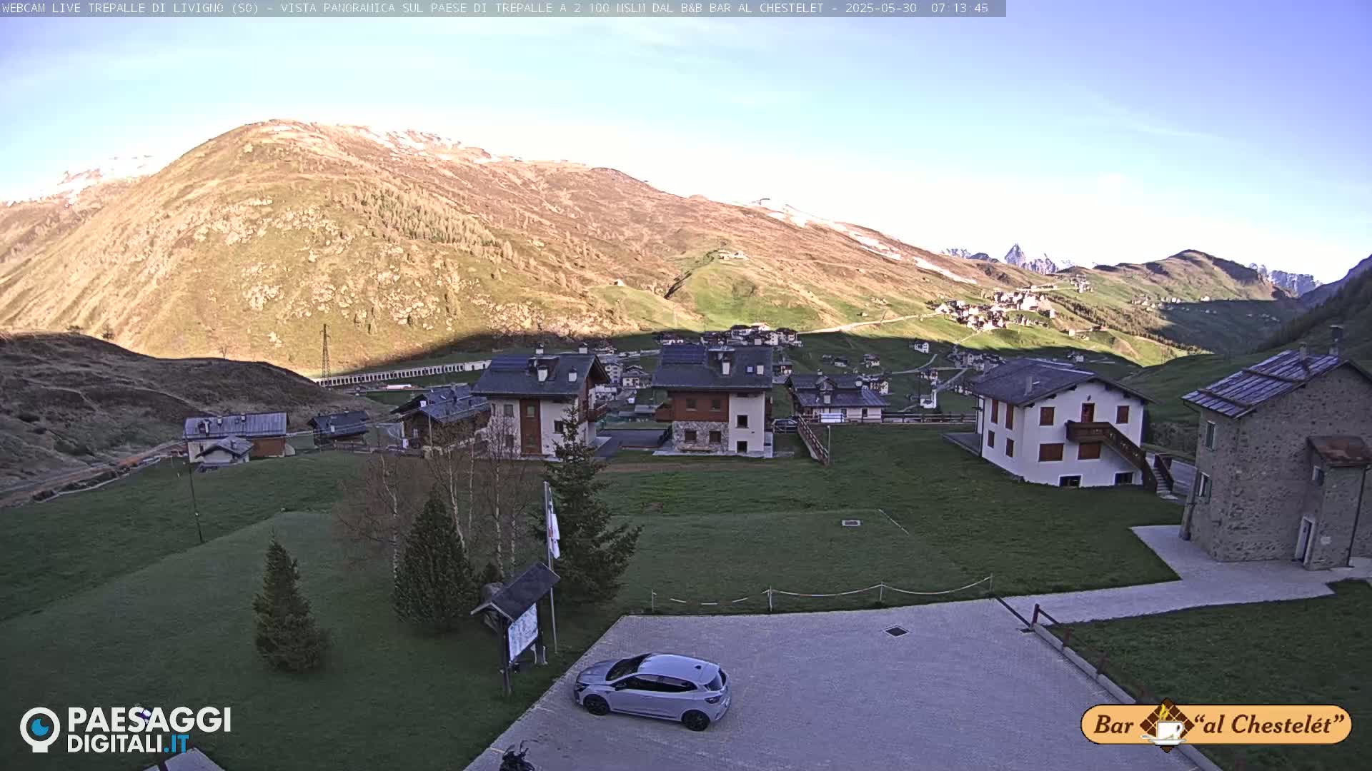 A sunny day reveals a small village nestled in a valley surrounded by mountains, with a car parked in a lot nearby.