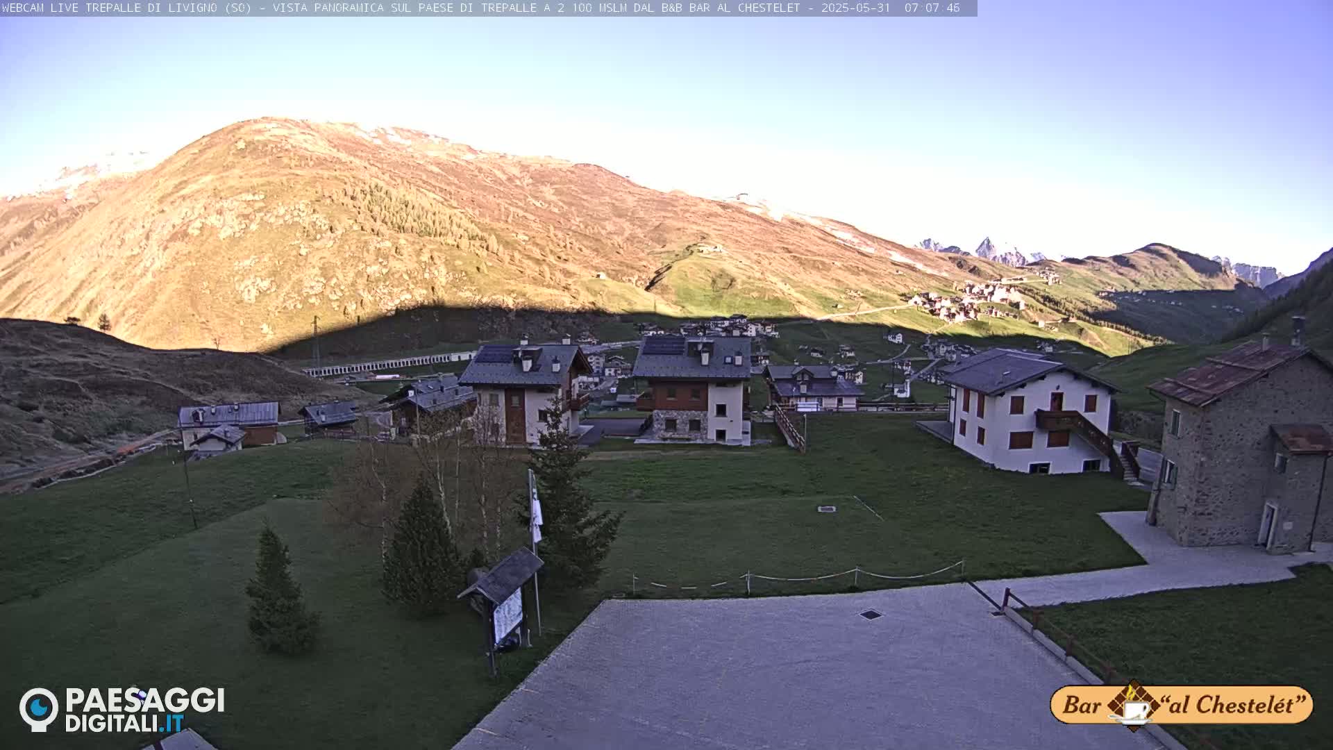 A sunny morning view of a small village nestled in a mountainous valley, with several buildings and grassy areas visible.