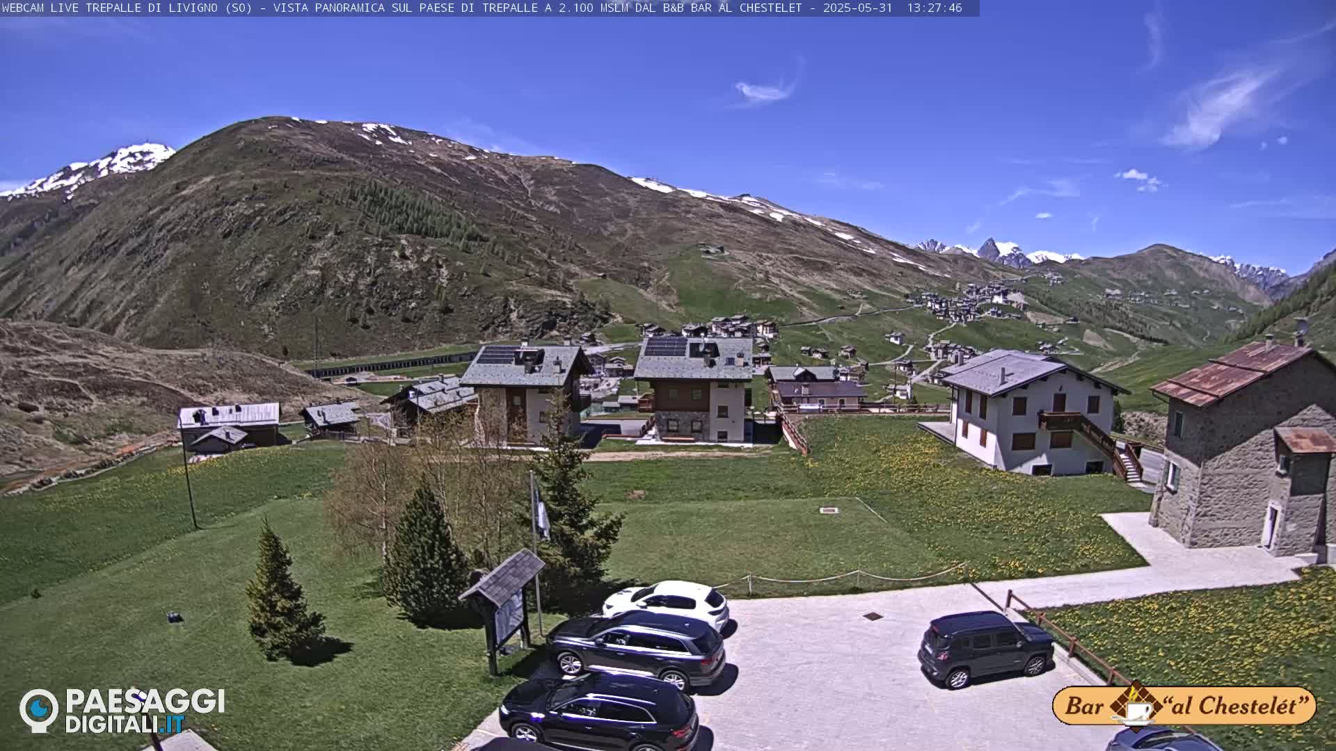 A sunny mountain village with a parking lot of several cars is nestled below a snow-capped mountain range.