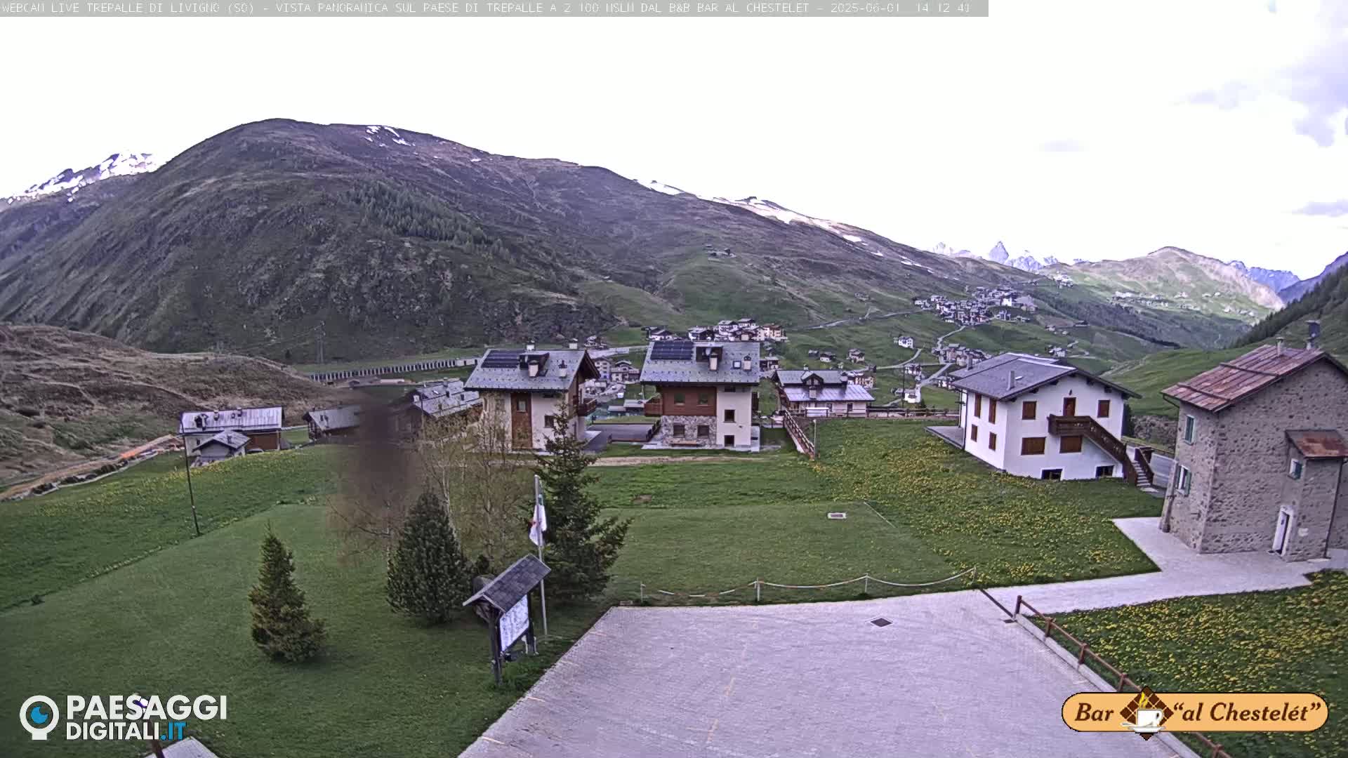 A small village nestled in a green valley, surrounded by mountains with patches of snow, under a mostly clear sky.