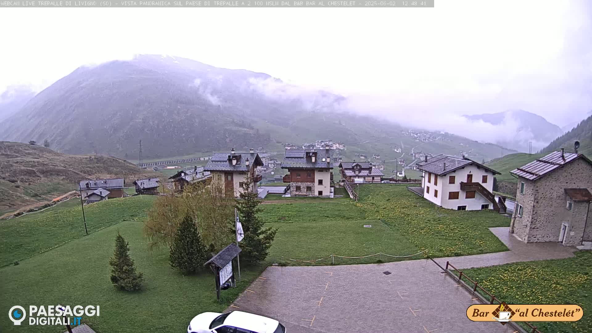 A misty mountain village with several buildings is seen from an elevated vantage point, with green grassy areas surrounding them.