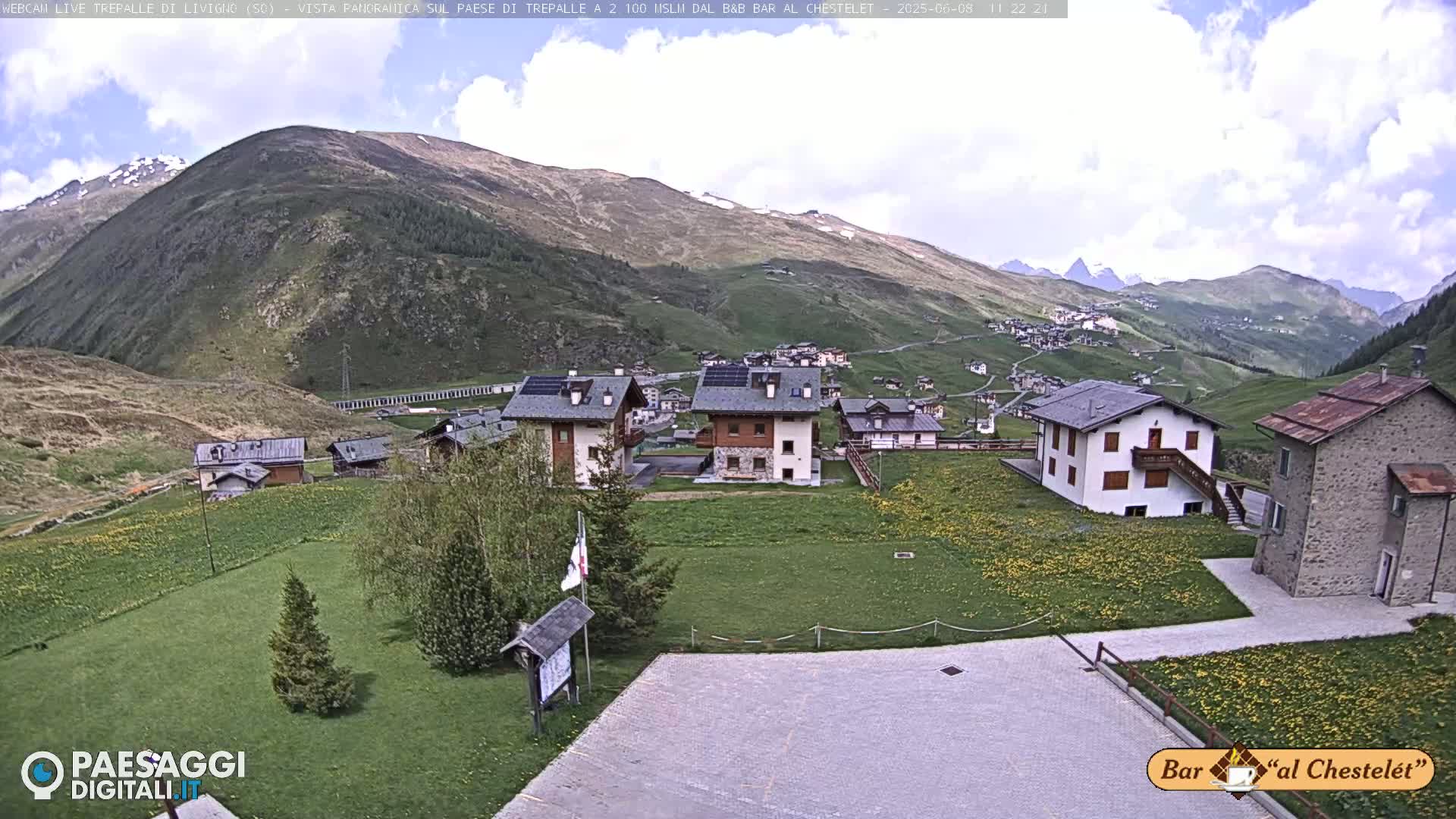A partly cloudy day reveals a village nestled in a valley surrounded by green hills and mountains, some with snow patches.