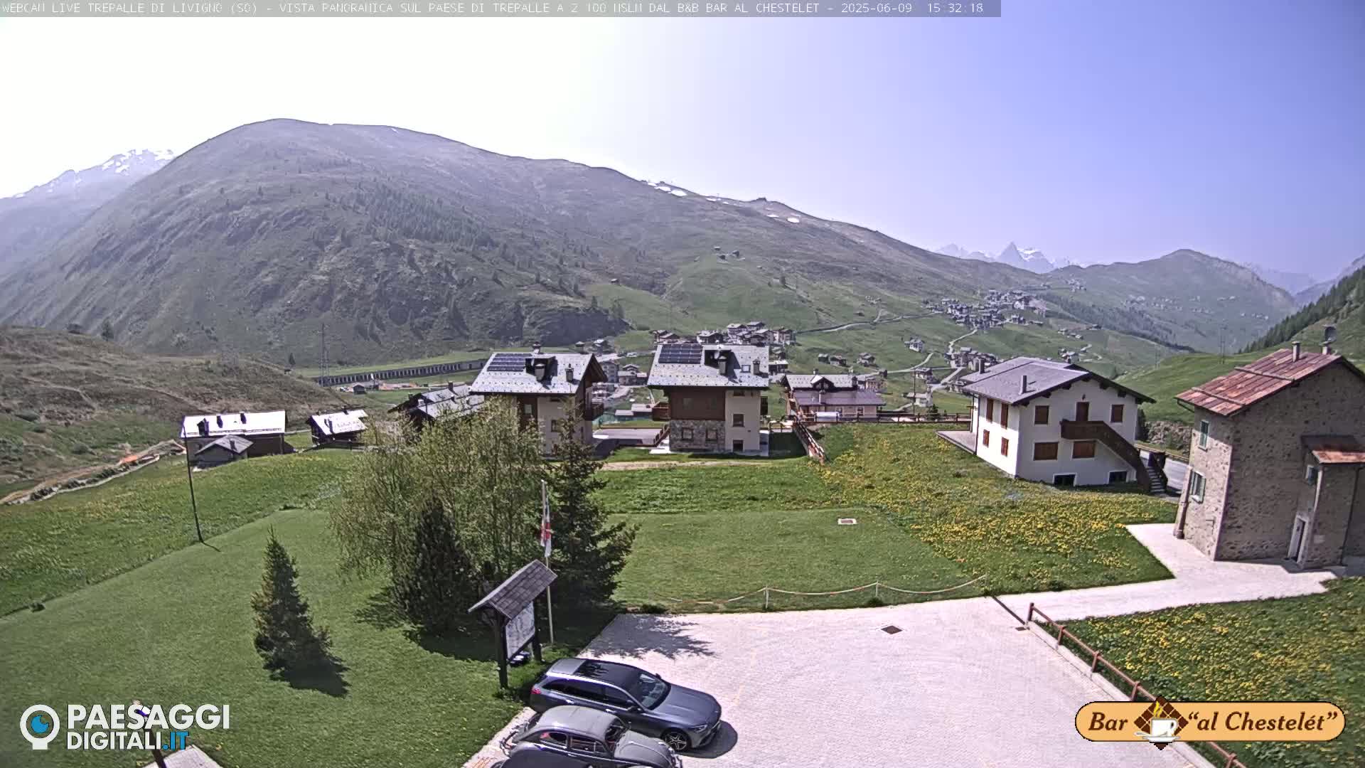 A sunny mountain village nestled in a valley, with several buildings and a few cars parked in a lot, overlooks a larger town further down the valley.