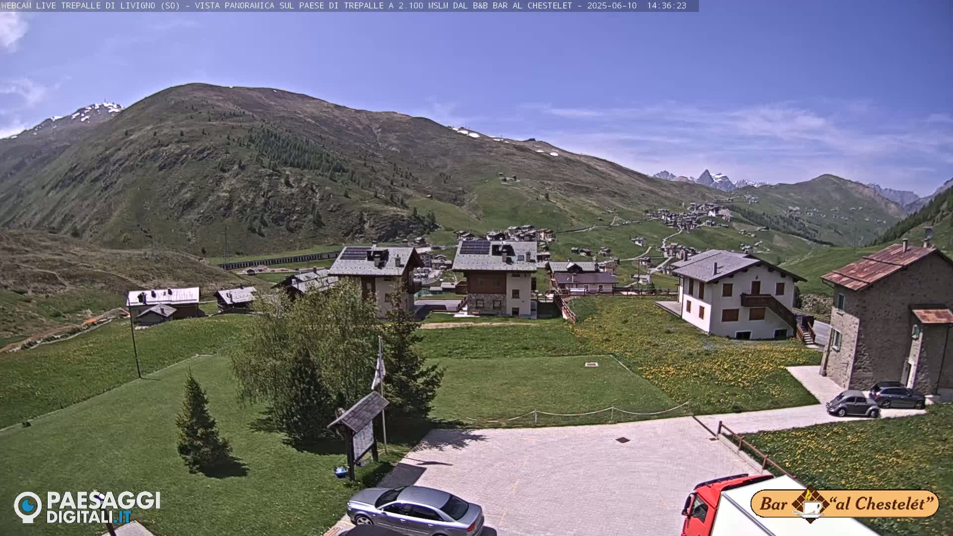 A sunny view of a village nestled in a mountainous valley, with a few cars parked in a lot and a truck in the foreground.
