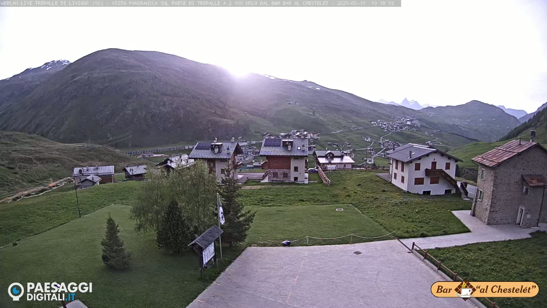 A mountain village with several buildings is nestled in a valley, bathed in the soft light of a setting sun.