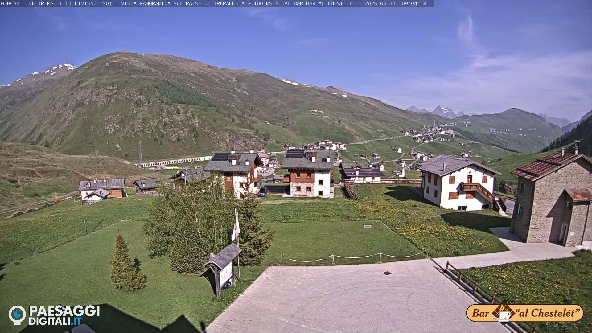 A sunny mountain village nestled in a green valley, with a few buildings and patches of grass, is overlooked by rolling hills and distant snow-capped peaks.