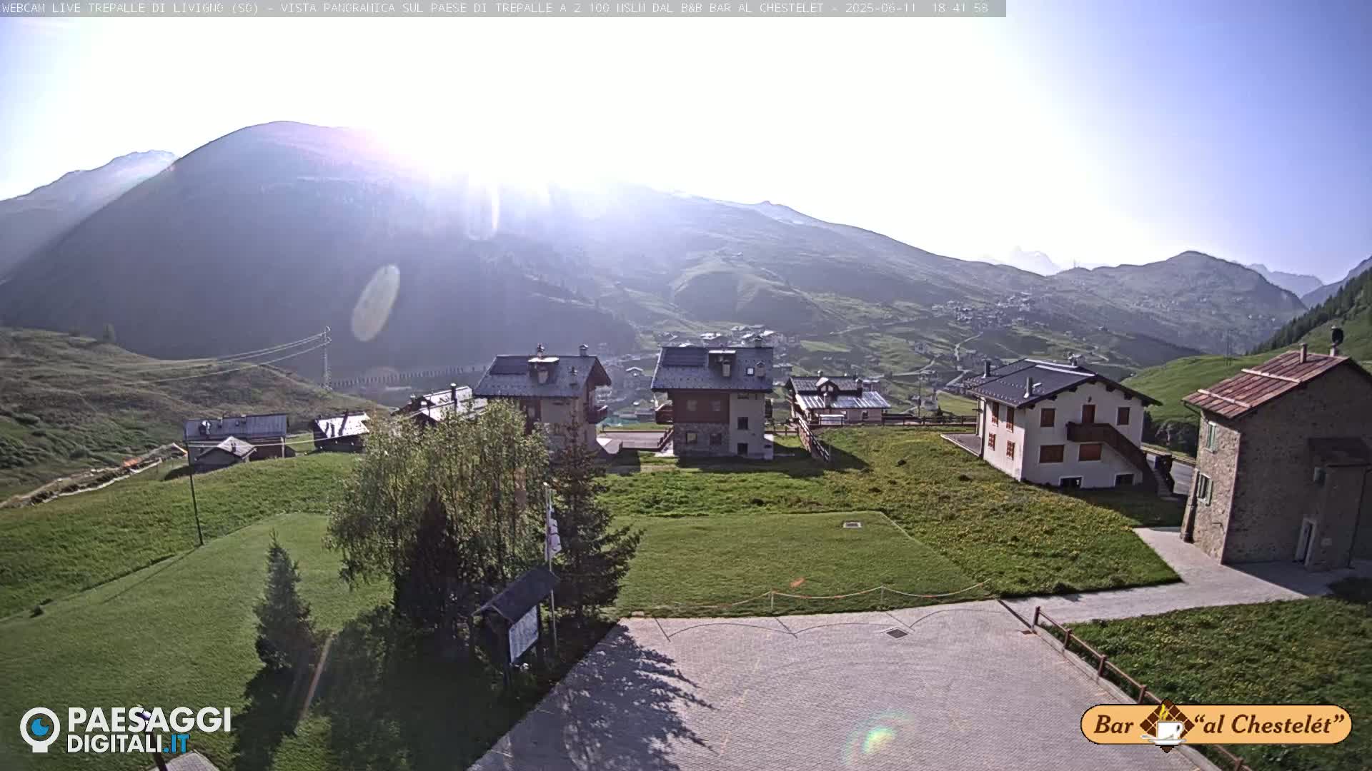 A sunny mountainside village scene shows several houses and grassy areas, with mountains in the background.