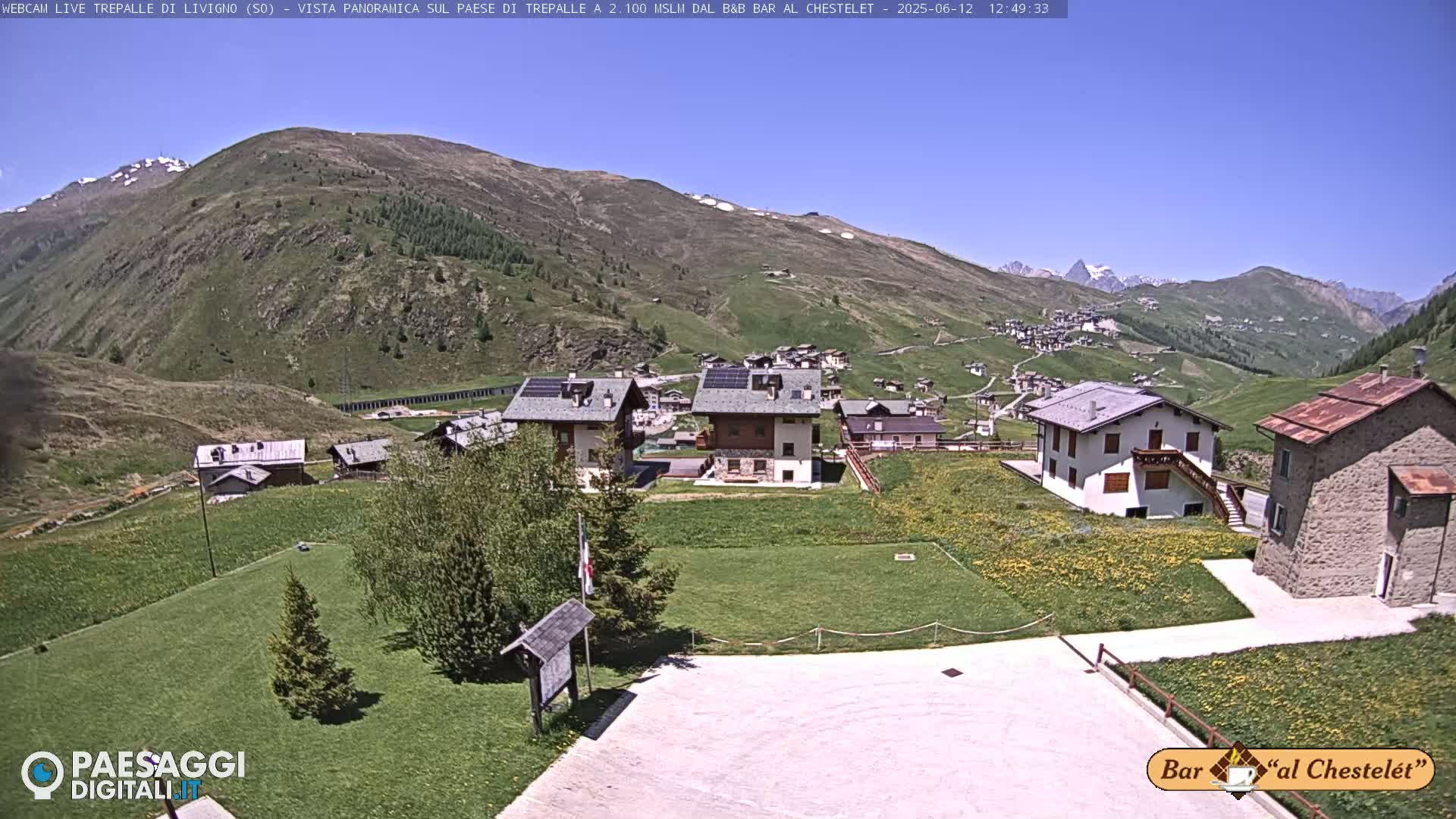 A sunny mountain village nestled in a valley, with green hills and a clear blue sky.