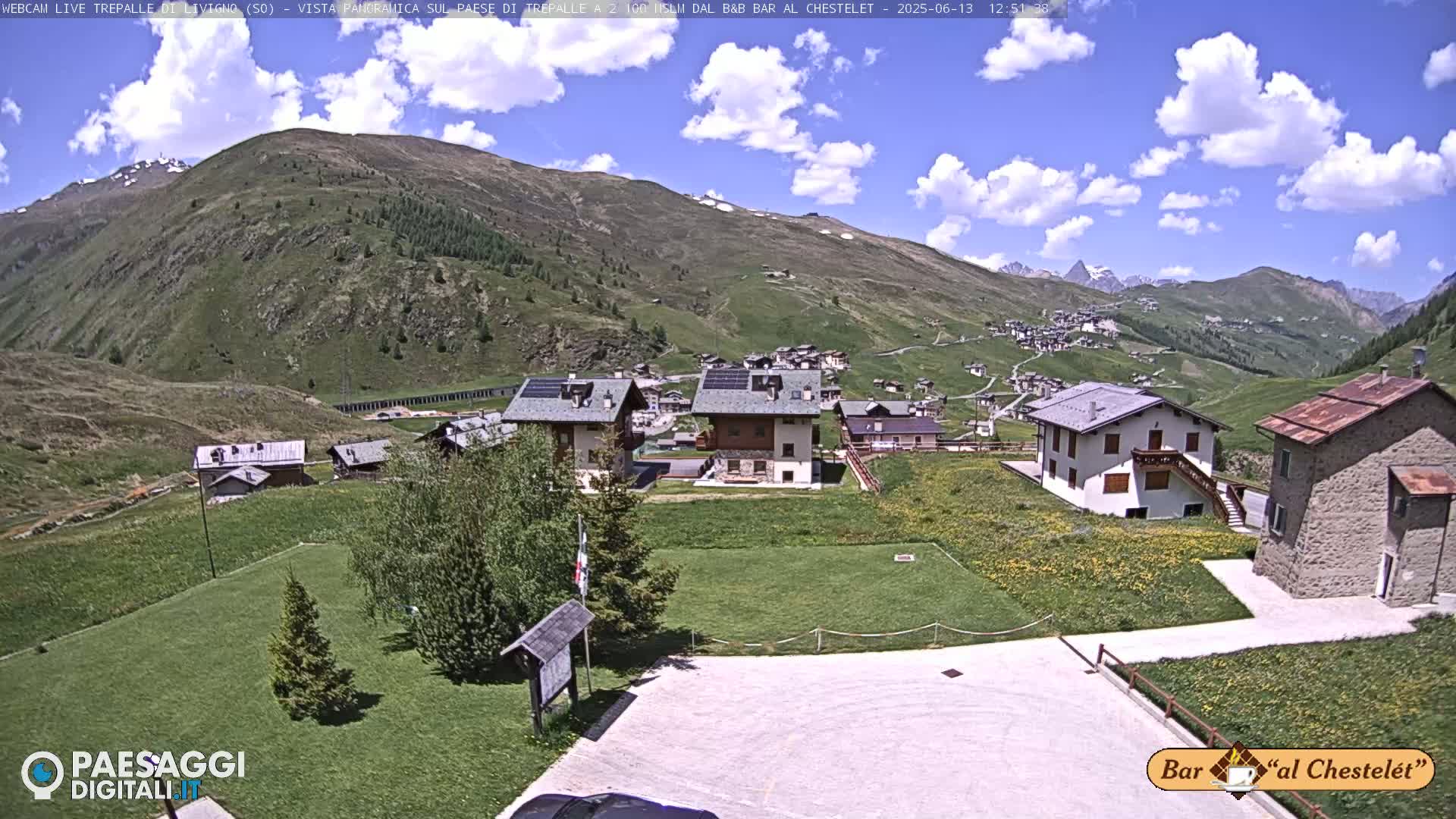A sunny day with partly cloudy skies shows a small village nestled in a mountain valley, with several buildings visible and green grassy areas surrounding them.