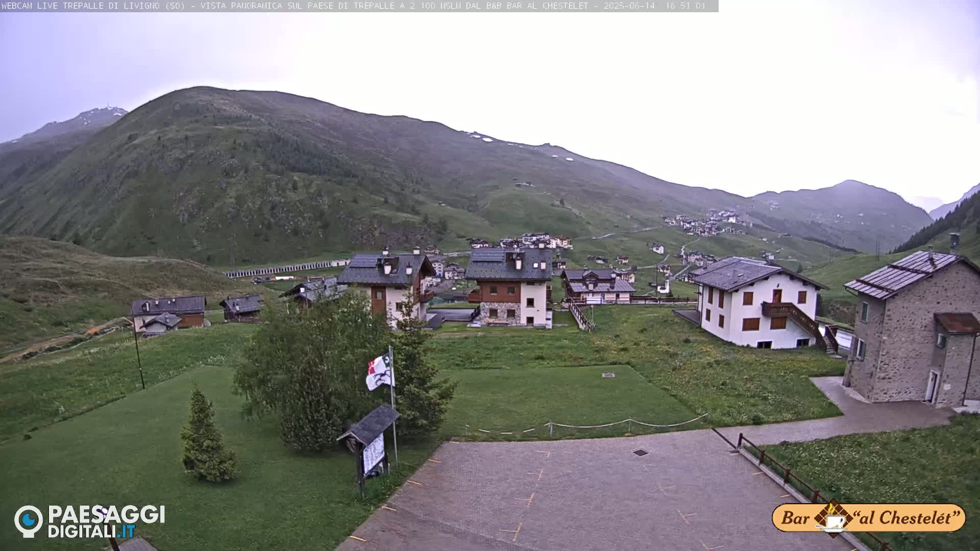 A small village nestled in a green valley is partially shrouded in overcast conditions, with mountains rising in the background.