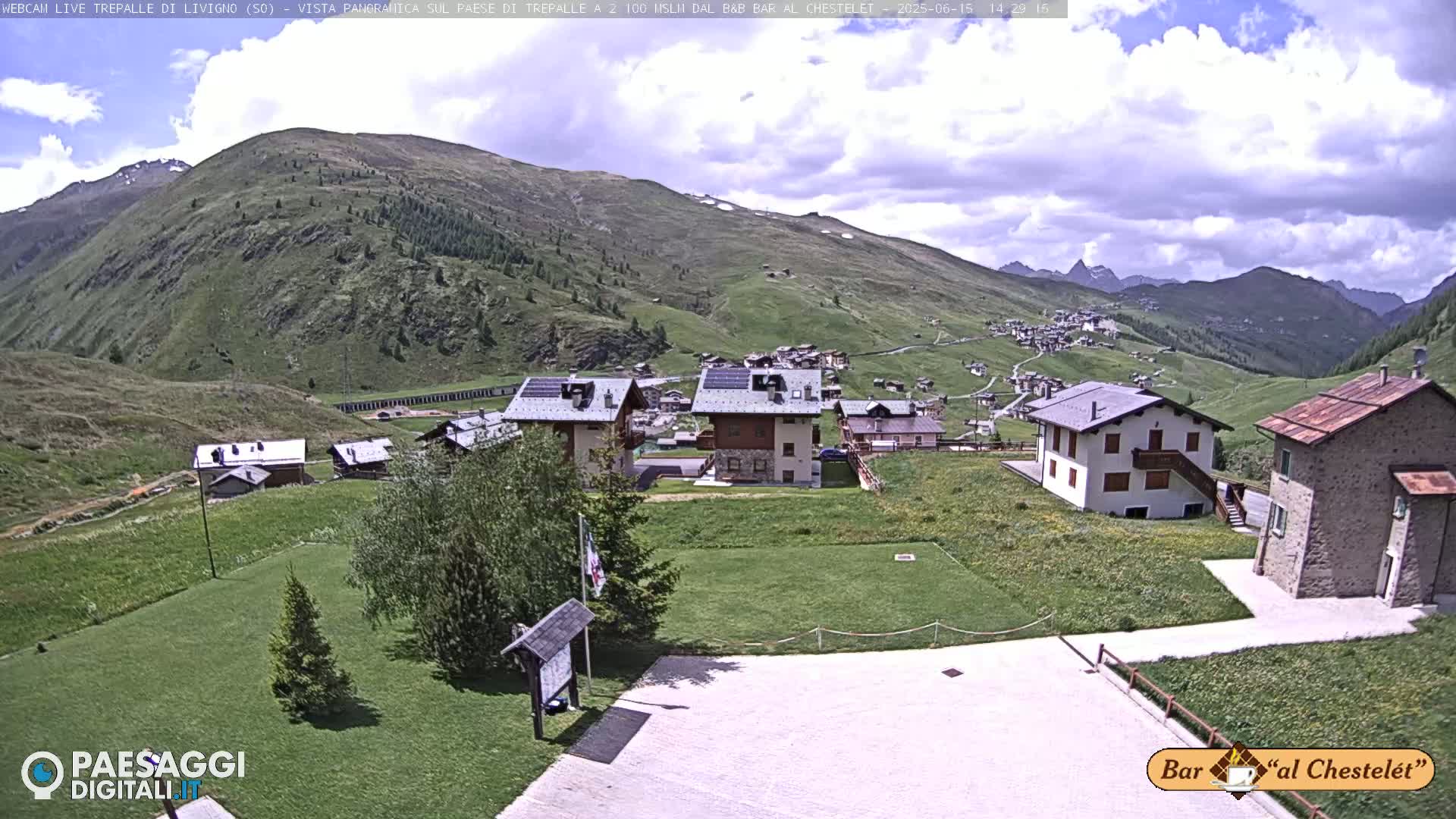 A partly cloudy day reveals a mountain village nestled in a green valley, with additional buildings and grassy areas in the foreground.