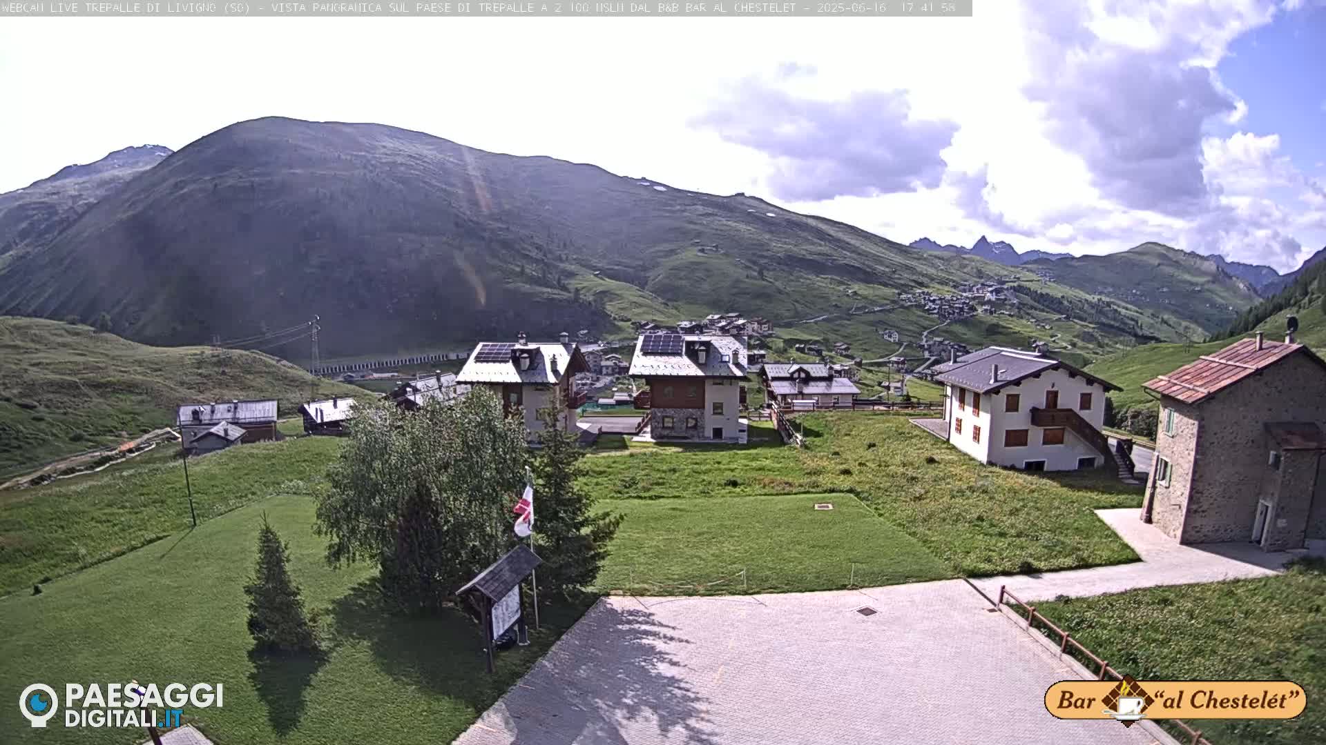 A partly cloudy day reveals a mountain village nestled in a valley, with several buildings and green grassy areas visible.