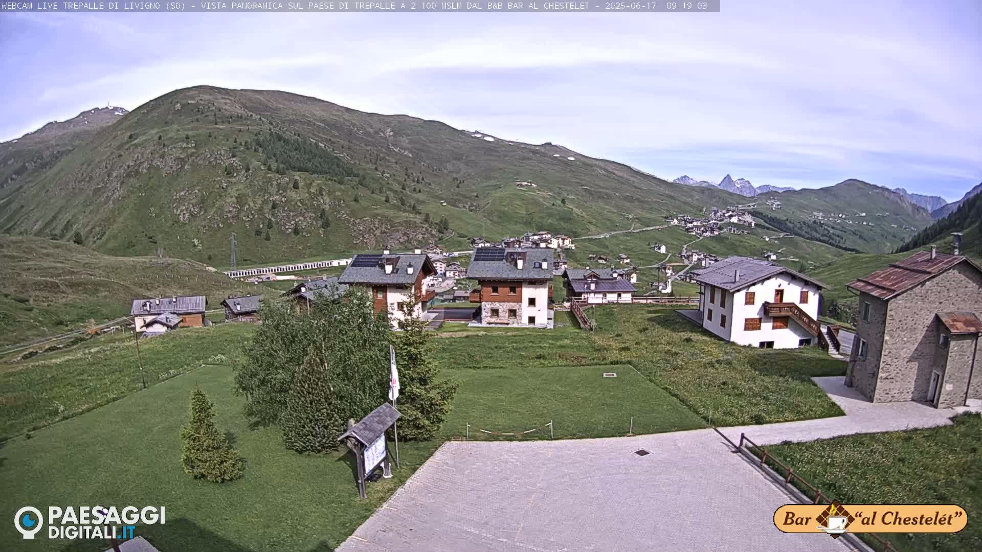 A mountain village nestled in a valley under a clear sky, surrounded by green hills and distant mountains.