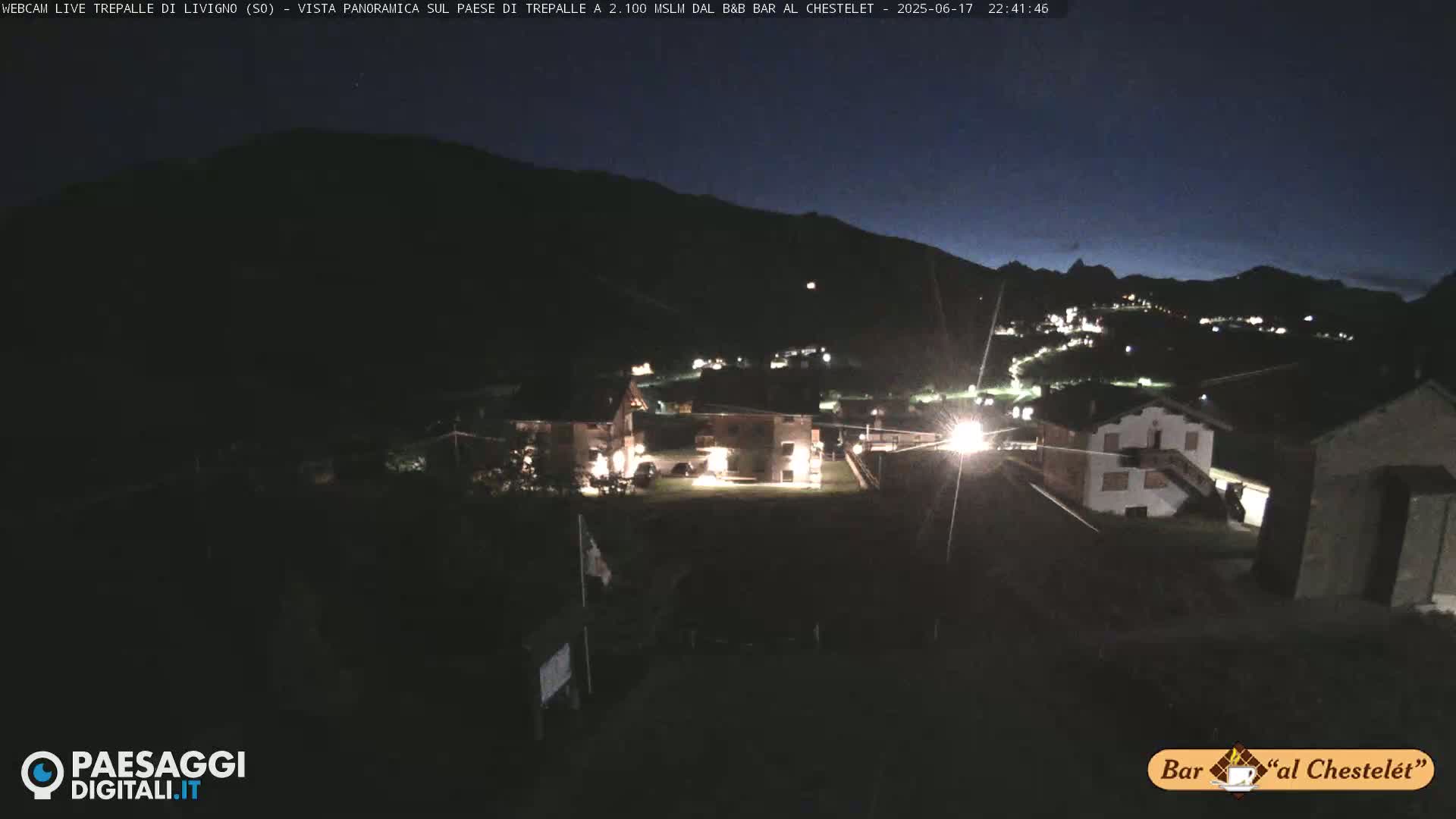 A nighttime, panoramic view of a small village nestled in a valley, surrounded by dark mountains under a clear, dark sky.