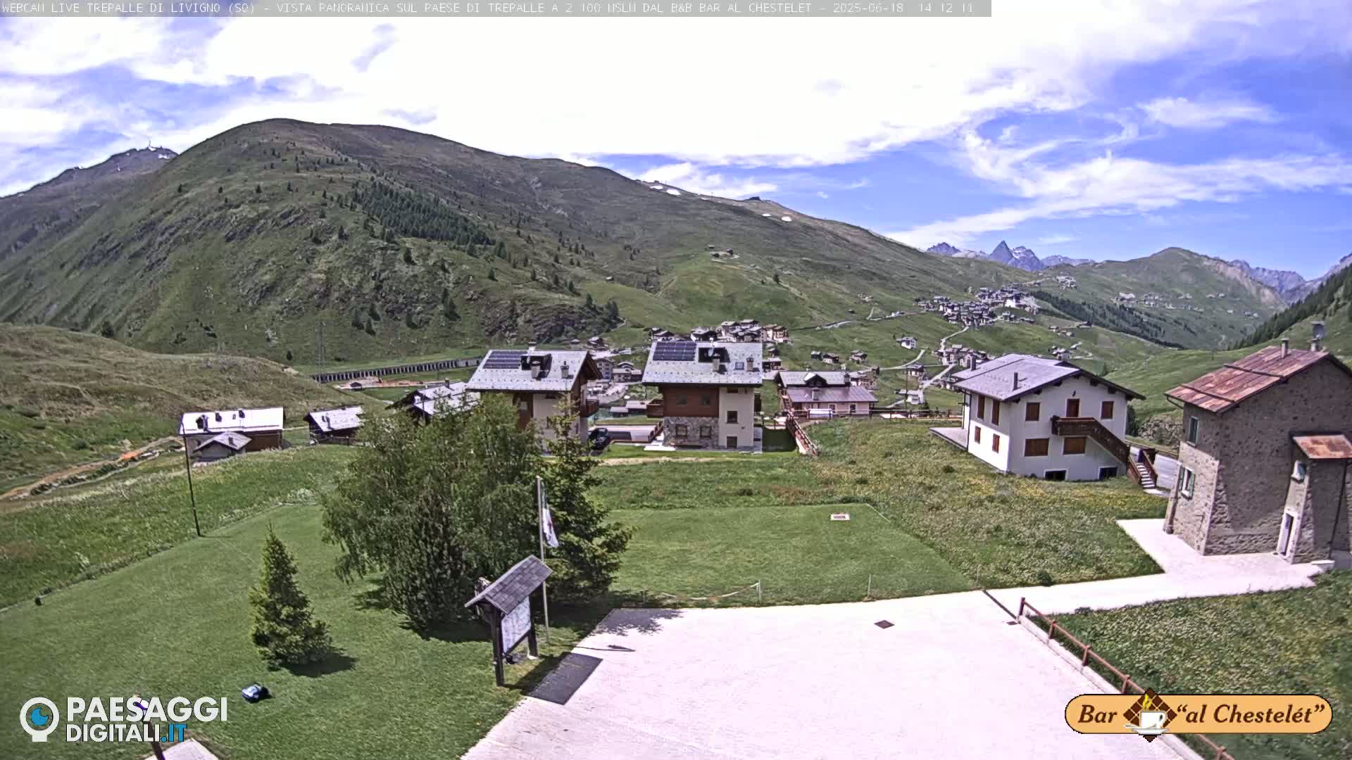 A sunny day reveals a mountain village nestled in a valley, with several houses and grassy areas visible, and mountains in the background.