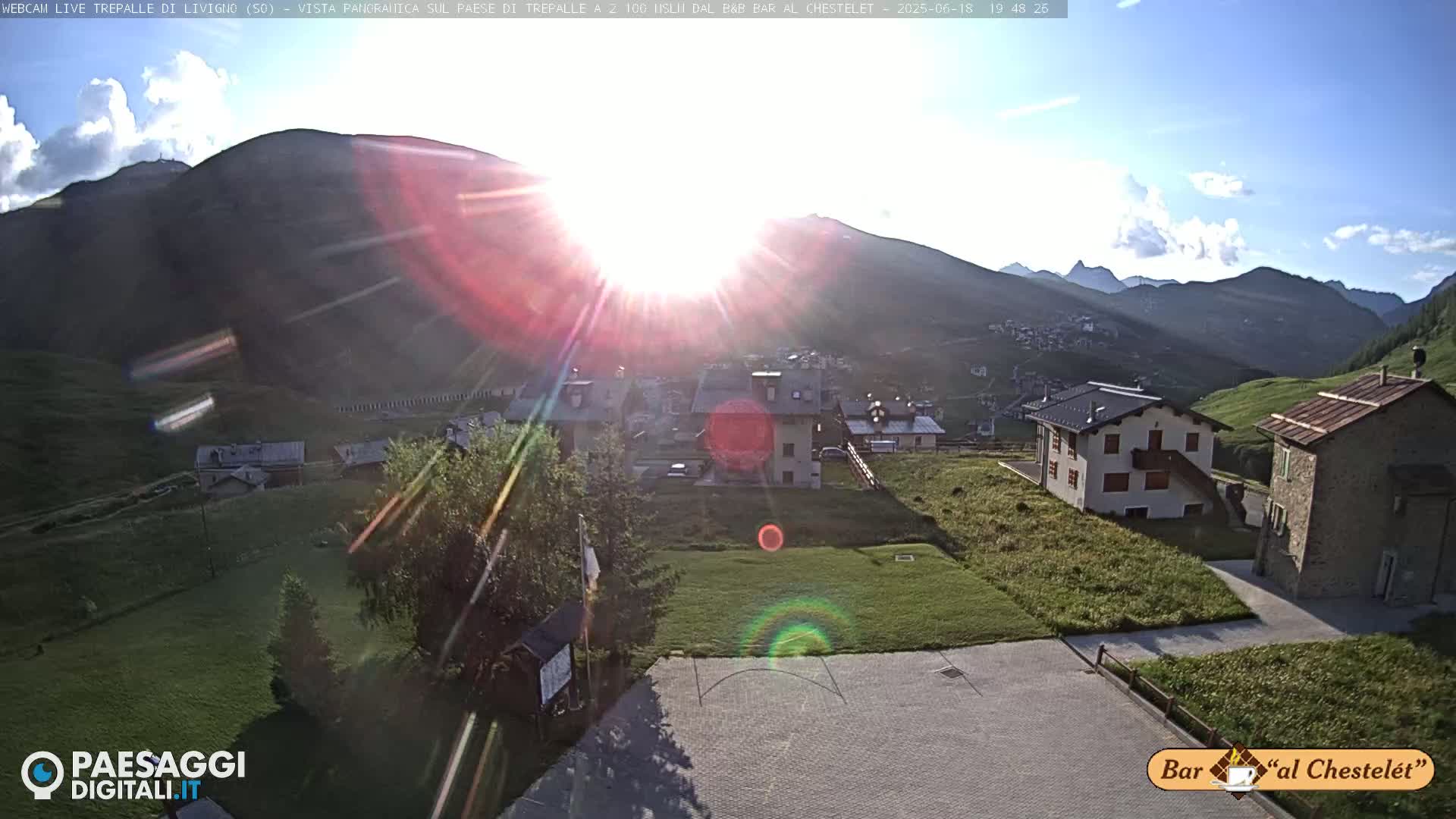 A sunny, high-altitude village nestled in a mountain valley, with several buildings visible and green grass surrounding a paved area.