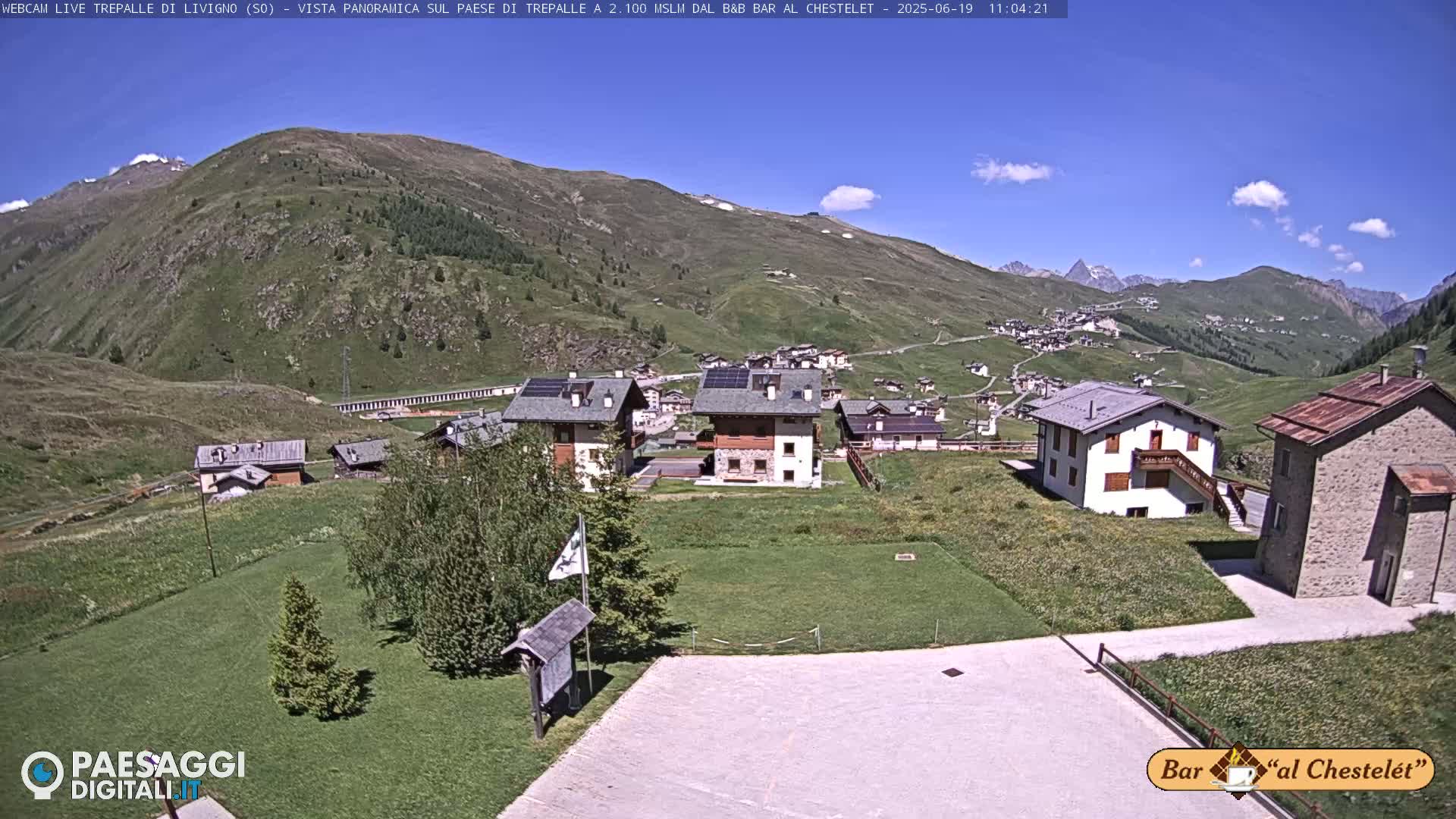 A sunny mountain village nestled in a valley, with several buildings and grassy areas, under a clear blue sky.