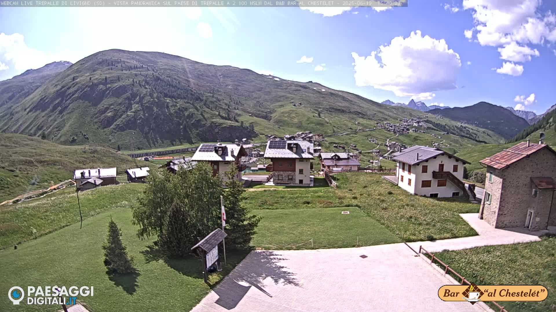 A sunny day reveals a mountainous village scene with several buildings, grassy areas, and a paved parking lot.