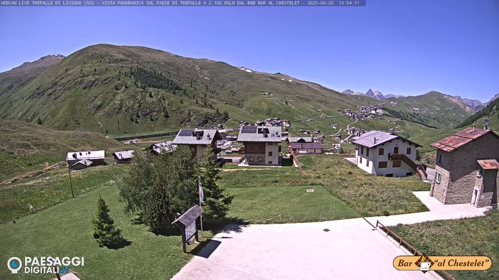 A sunny day reveals a mountain village nestled in a valley, with several buildings visible and grassy areas surrounding them.
