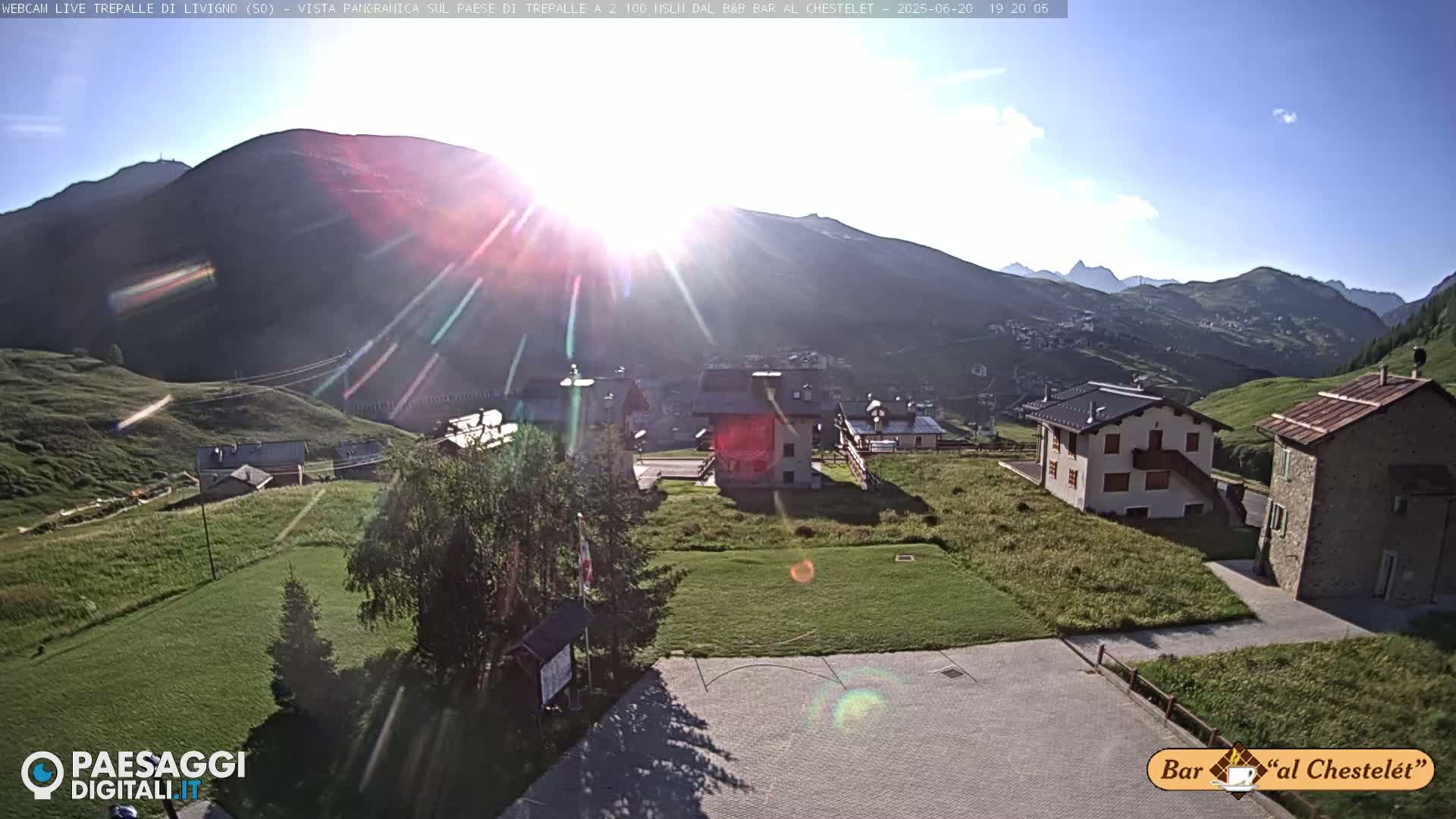 A sunny mountain vista shows several buildings in a small village, with green grass and a paved area in the foreground.